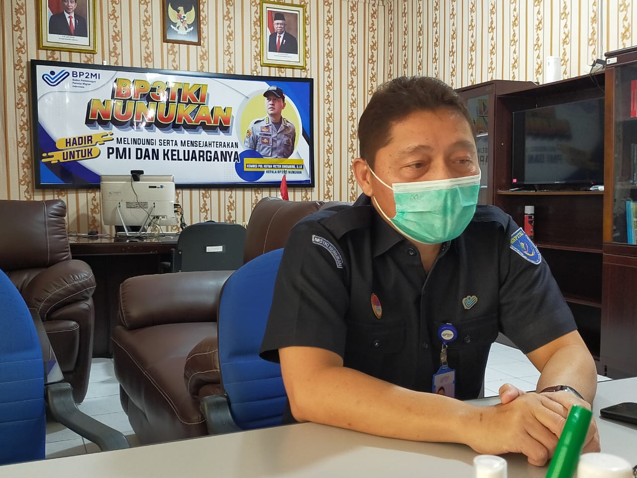  The image shows a man wearing a surgical mask and a navy uniform with the logo of the Indonesian Ministry of Manpower. He is sitting at a desk in an office, with a computer and a television in the background.